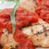 Grilled chicken breast topped with chunky fresh tomato tarragon sauce and a green tarragon leaf on a white plate.