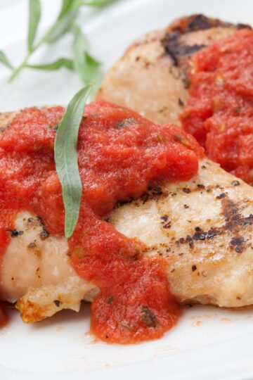 Grilled chicken breast topped with chunky fresh tomato tarragon sauce and a green tarragon leaf on a white plate.