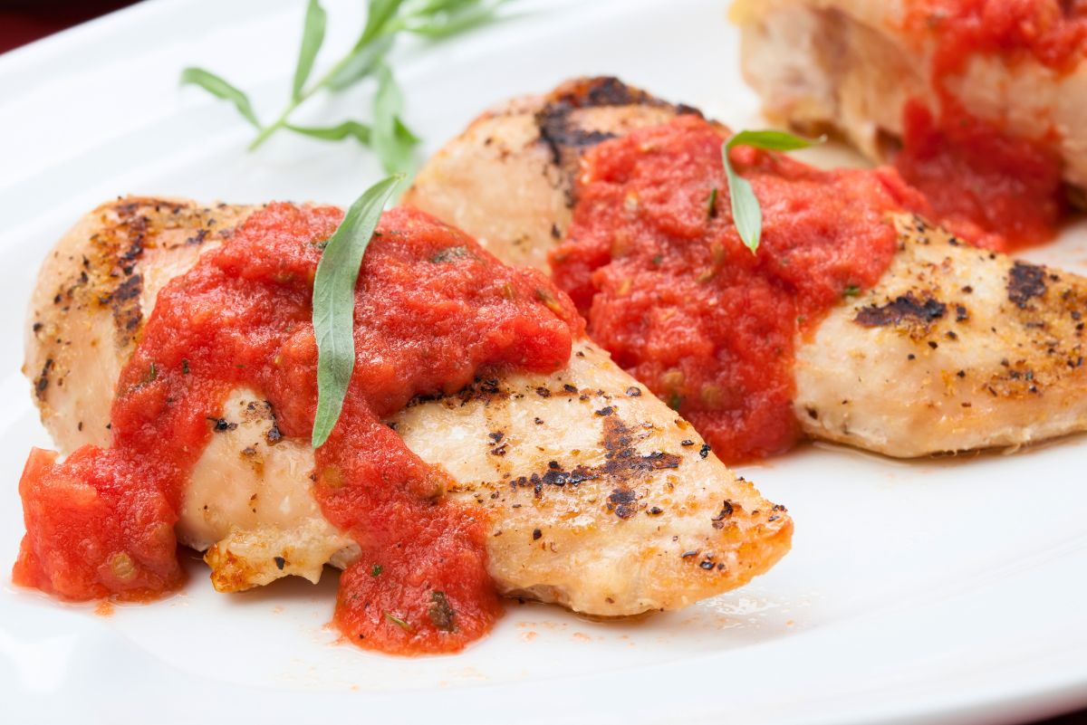 Grilled chicken breasts topped with fresh tomato tarragon sauce and green tarragon leaves on a white plate.