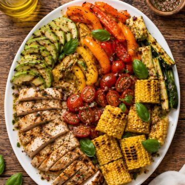 Grilled corn, chicken and summer vegetable salad with sliced grilled chicken, corn on the cob, zucchini, bell peppers, tomatoes, avocado, basil, and vinaigrette on a white platter.