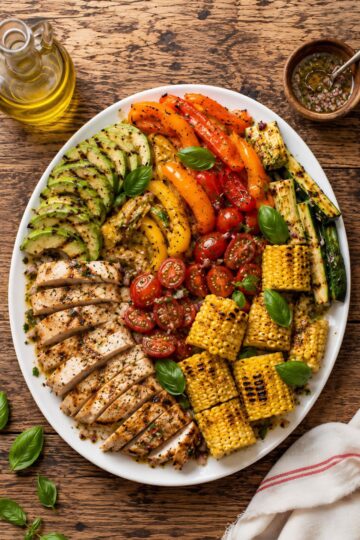 Grilled corn, chicken and summer vegetable salad with sliced grilled chicken, corn on the cob, zucchini, bell peppers, tomatoes, avocado, basil, and vinaigrette on a white platter.