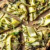 Close-up of grilled pesto zucchini pizza slices with melted cheese, curled zucchini ribbons, basil, red pepper flakes, and charred crust on a wooden board.