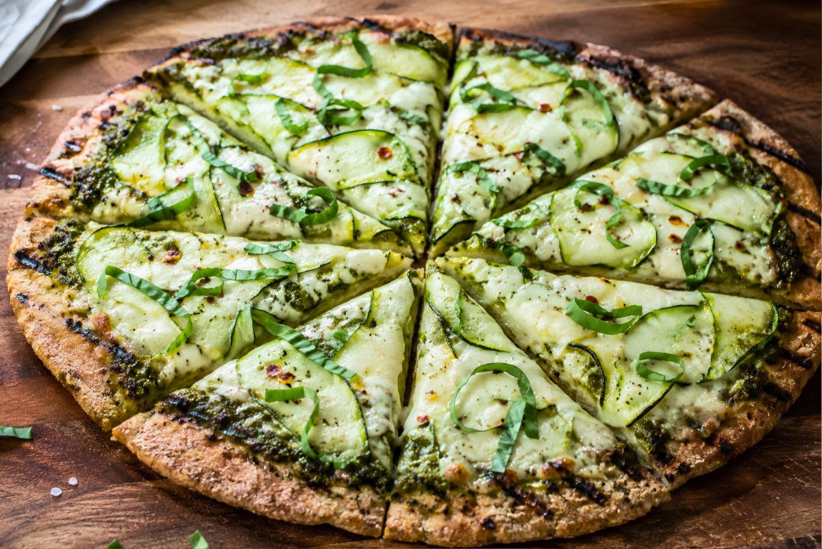 Whole grilled pesto zucchini pizza sliced on a wooden board with melted mozzarella, zucchini ribbons, fresh basil, red pepper flakes, and a charred golden crust.
