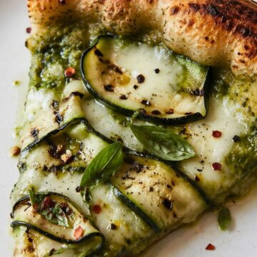 Slice of grilled pesto zucchini pizza on a white plate with charred crust, melted mozzarella, zucchini ribbons, basil leaves, black pepper, and red pepper flakes.