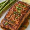 Grilled teriyaki salmon fillet with a glossy glaze, sesame seeds, and green onion on a plate with roasted asparagus and a lemon wedge.