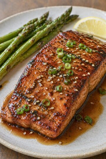 Grilled teriyaki salmon fillet with a glossy glaze, sesame seeds, and green onion on a plate with roasted asparagus and a lemon wedge.