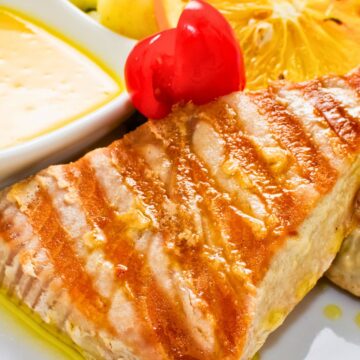 Grilled tuna steak with golden-brown sear marks on a white plate, topped with a glossy red cherry tomato. Served with a lightly grilled orange slice, sautéed zucchini and potatoes, and a small dish of creamy lemon butter sauce. Drizzled olive oil adds a glistening finish.