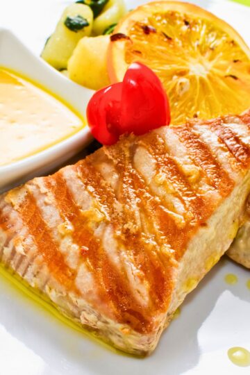 Grilled tuna steak with golden-brown sear marks on a white plate, topped with a glossy red cherry tomato. Served with a lightly grilled orange slice, sautéed zucchini and potatoes, and a small dish of creamy lemon butter sauce. Drizzled olive oil adds a glistening finish.