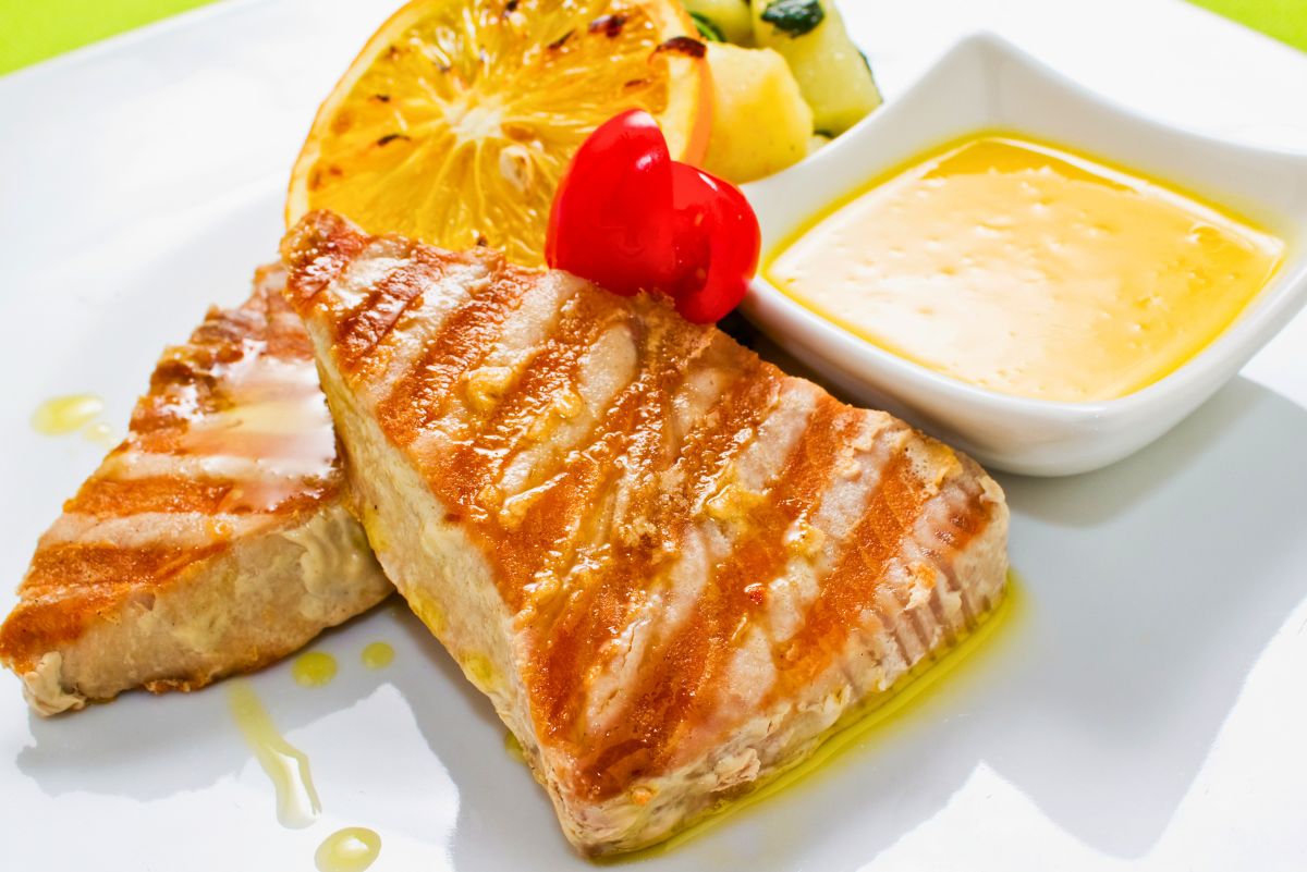 Grilled tuna steaks with golden-brown sear marks on a white plate, served with a small dish of creamy lemon butter sauce. Accompanied by a lightly grilled orange slice, sautéed zucchini and potatoes, and a bright red cherry tomato garnish, with drizzled olive oil adding a glistening finish.