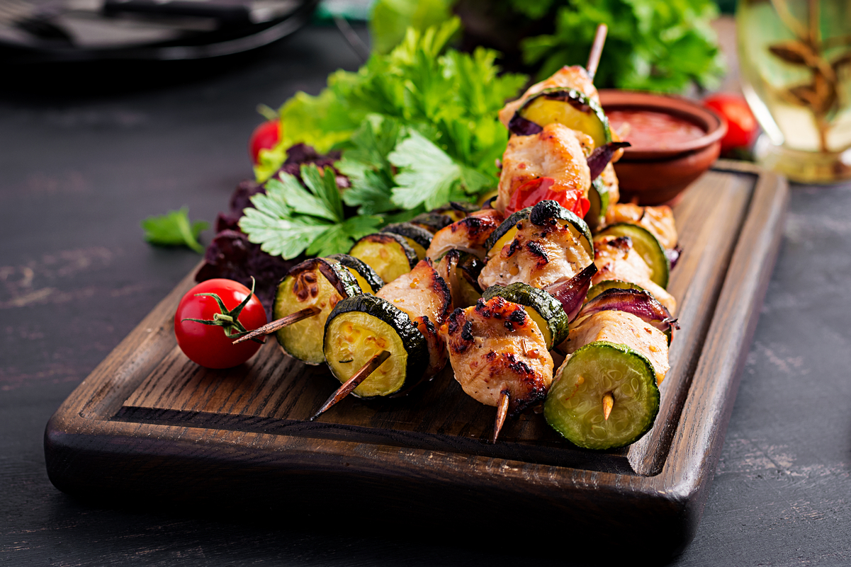 Grilled Moroccan chicken kebabs with zucchini, red onion, and chicken breast on skewers, served on a wooden board with fresh herbs, cherry tomatoes, and a small bowl of sauce.