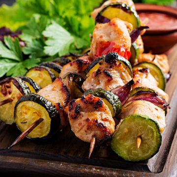 Grilled Moroccan chicken kebabs with zucchini, red onion, and cherry tomatoes on skewers, served on a wooden board with fresh herbs and a small bowl of sauce.
