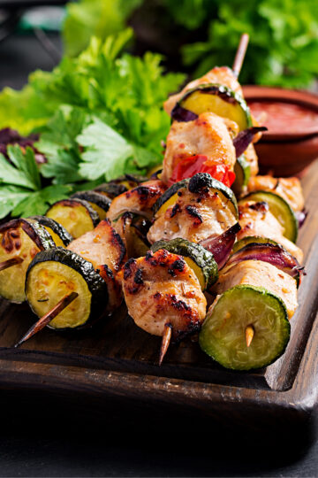 Grilled Moroccan chicken kebabs with zucchini, red onion, and cherry tomatoes on skewers, served on a wooden board with fresh herbs and a small bowl of sauce.