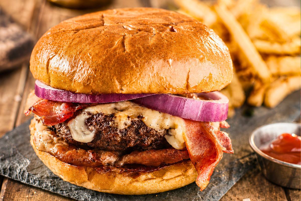 High protein bacon cheeseburger with melted cheese, crispy bacon, red onion, and a glossy toasted bun, served on a dark plate with fries and ketchup in the background.