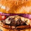 High protein bacon cheeseburger with a glossy toasted bun, melted cheese, crispy bacon, red onion, tomato, and a grilled beef patty on a dark plate.