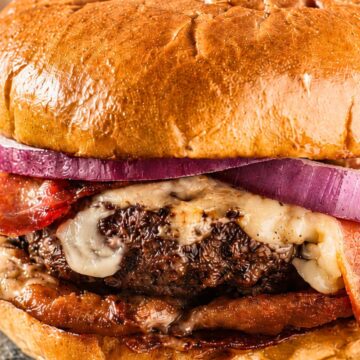 High protein bacon cheeseburger with a glossy toasted bun, melted cheese, crispy bacon, red onion, tomato, and a grilled beef patty on a dark plate.