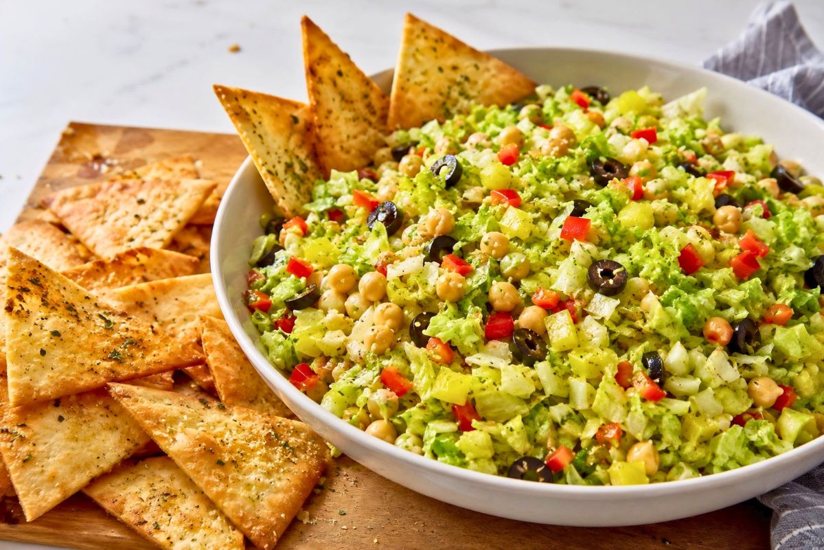 Italian chopped salad with garlic pita chips, chopped lettuce, chickpeas, black olives, banana peppers, red bell pepper, and herbs in a white bowl beside crispy pita chips.