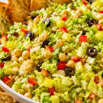 Italian chopped salad with garlic pita chips, chopped lettuce, chickpeas, black olives, banana peppers, red bell pepper, and herbs in a white bowl on a wooden surface.