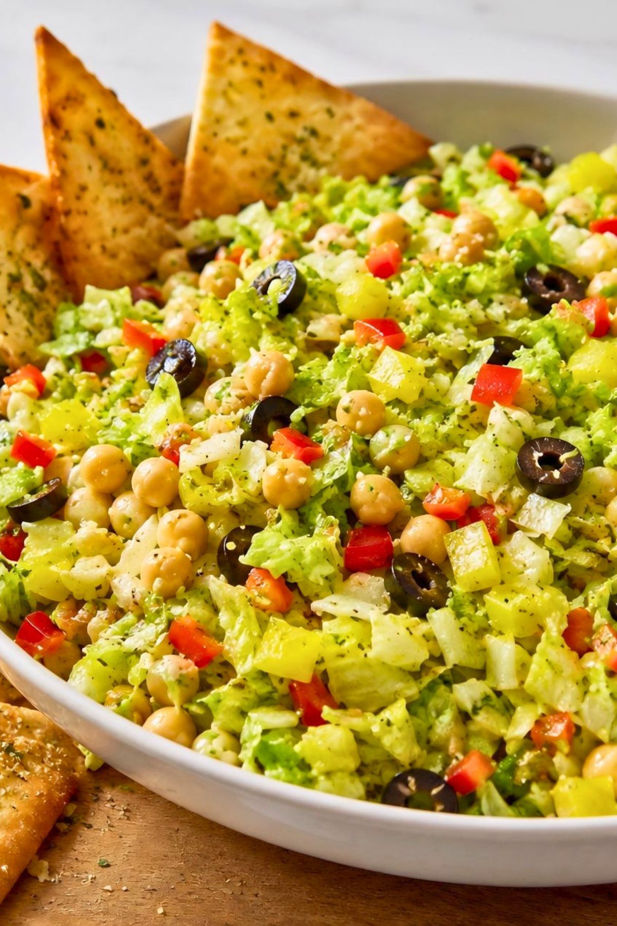 Italian chopped salad with garlic pita chips, chopped lettuce, chickpeas, black olives, banana peppers, red bell pepper, and herbs in a white bowl on a wooden surface.