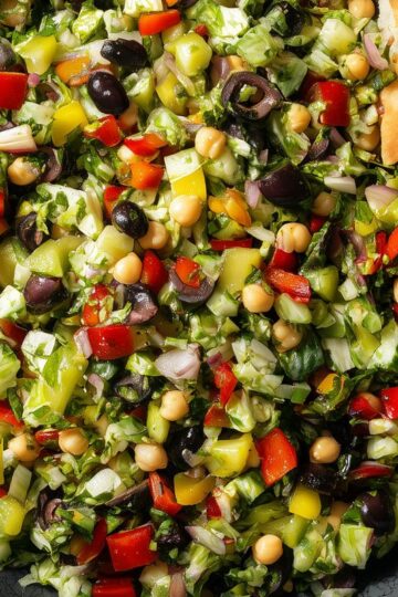 Italian chopped salad with finely chopped lettuce, chickpeas, red bell peppers, black olives, banana peppers, red onion, and garlic pita chips in a black bowl.
