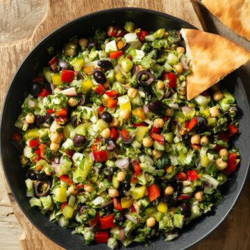 Italian chopped salad with lettuce, chickpeas, bell peppers, black olives, banana peppers, and red onion in a black bowl on a wooden cutting board with garlic pita chips.