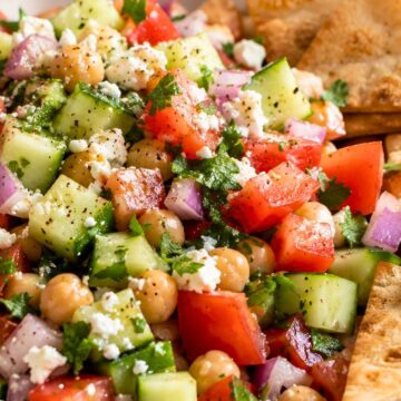 Mediterranean cucumber tomato chopped salad with chickpeas, feta cheese, red onion, herbs, and black pepper in a bowl with crispy pita chips on the side.