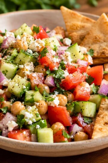 Mediterranean cucumber tomato chopped salad with chickpeas, feta cheese, red onion, herbs, and black pepper in a bowl with crispy pita chips on the side.