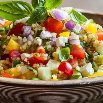 Mediterranean quinoa salad with cucumber, tomato, yellow bell pepper, red onion, feta cheese, and fresh basil piled in a rustic bowl.