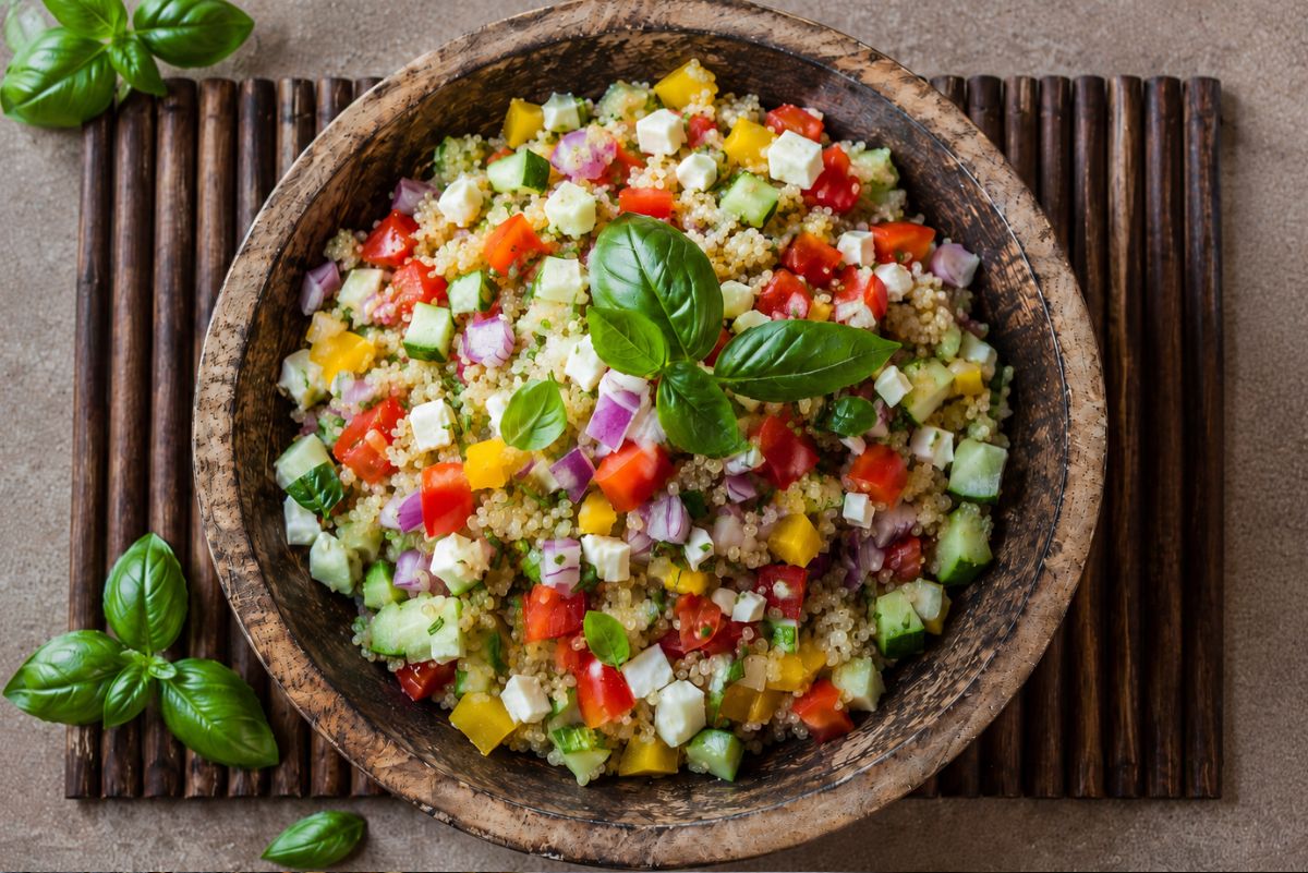Mediterranean quinoa salad with cucumber, tomato, yellow bell pepper, red onion, feta cheese, and fresh basil in a rustic wooden bowl on a dark bamboo tray.