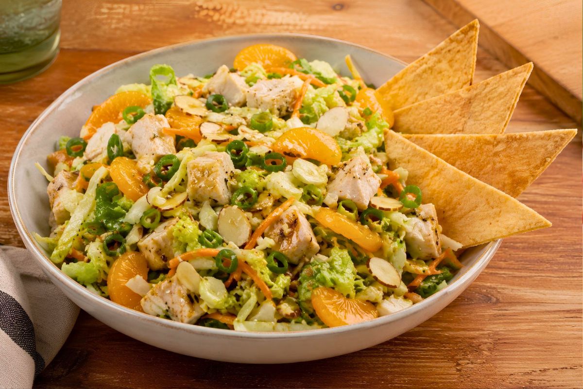 Sesame chicken chopped salad in a shallow bowl with diced chicken, mandarin oranges, romaine, cabbage, carrots, sliced almonds, green onions, and crispy wonton chips on a wooden table.