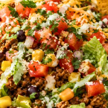 Close-up of a high-protein street taco chopped salad in a bowl. Features chopped romaine lettuce, seasoned ground beef, black beans, corn, diced tomatoes, shredded cheese, cilantro, and a creamy dressing drizzle. Tortilla chips are visible on the side, emphasizing a fresh and colorful Tex-Mex style salad.