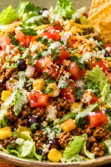 Close-up of a high-protein street taco chopped salad in a bowl. Features chopped romaine lettuce, seasoned ground beef, black beans, corn, diced tomatoes, shredded cheese, cilantro, and a creamy dressing drizzle. Tortilla chips are visible on the side, emphasizing a fresh and colorful Tex-Mex style salad.