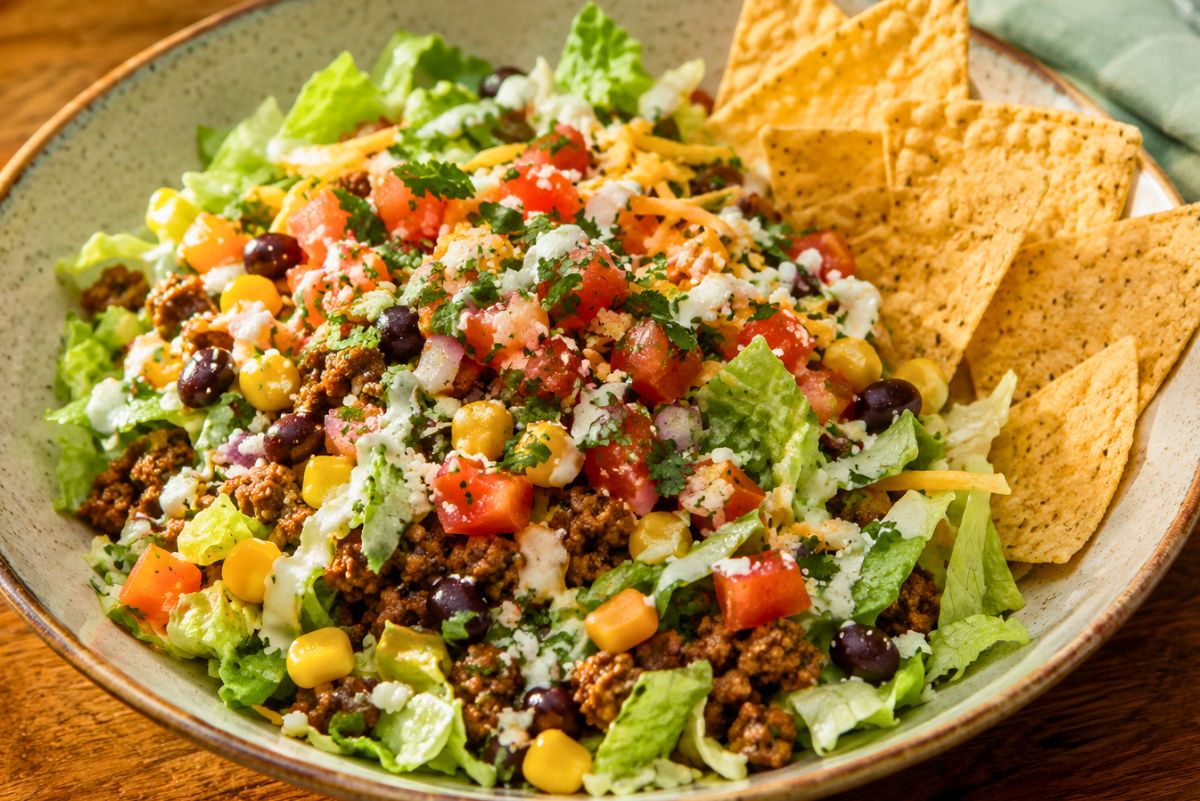 A colorful, high-protein street taco chopped salad in a ceramic bowl. The salad includes chopped romaine, seasoned ground beef, black beans, corn, diced tomatoes, shredded cheese, and fresh cilantro, drizzled with a creamy dressing. Tortilla chips are arranged on the side, highlighting a fresh, Tex-Mex style presentation.