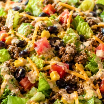 Taco chopped salad in a bowl with romaine lettuce, seasoned ground beef, black beans, corn, tomatoes, shredded cheddar cheese, black olives, and creamy taco ranch dressing.