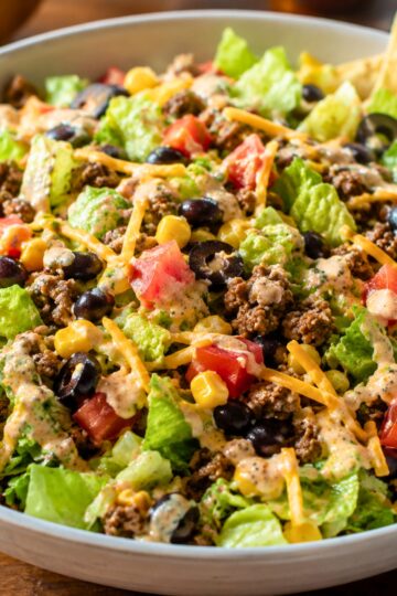 Taco chopped salad in a bowl with romaine lettuce, seasoned ground beef, black beans, corn, tomatoes, shredded cheddar cheese, black olives, and creamy taco ranch dressing.