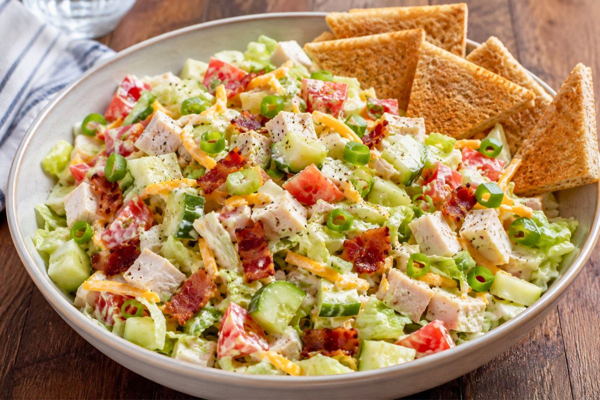 Turkey club chopped salad with toast chips, diced turkey, bacon, lettuce, tomato, cucumber, cheddar cheese, green onions, and creamy ranch dressing in a large bowl on a wooden table.