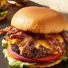 Close-up of a bacon cheeseburger with melted cheddar cheese, crispy bacon, lettuce, tomato, and red onion on a soft bun.