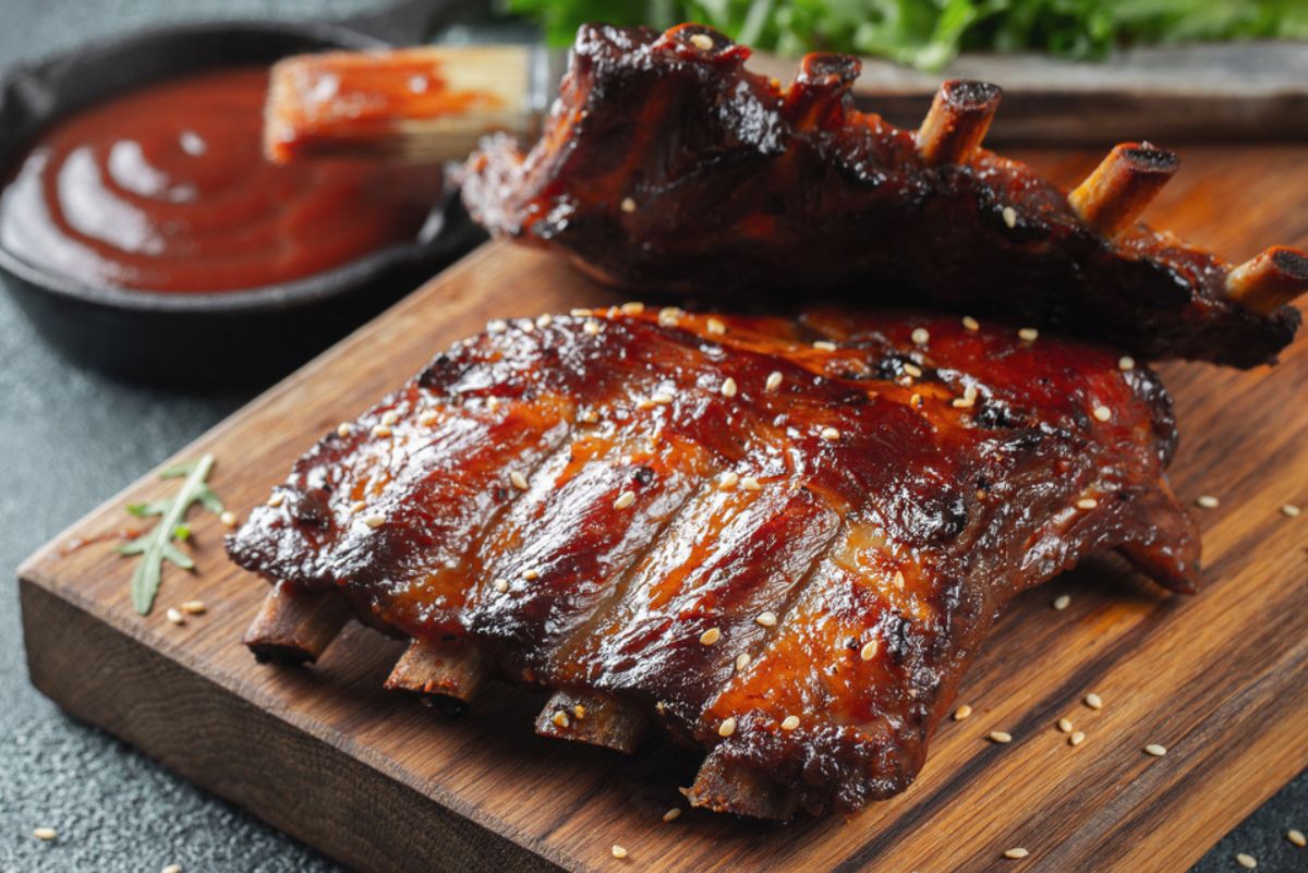 BBQ ribs with glossy sauce and sesame seeds on a wooden cutting board, served with barbecue sauce and a basting brush in the background.