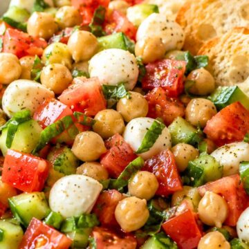 Caprese chickpea salad with chickpeas, mozzarella, tomatoes, cucumber, fresh basil, black pepper, and toasted baguette chips in a shallow bowl.