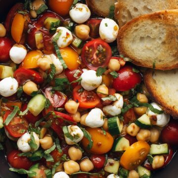 Close-up of Caprese chickpea salad with cherry tomatoes, mozzarella pearls, cucumbers, basil, balsamic dressing, and toasted baguette slices.