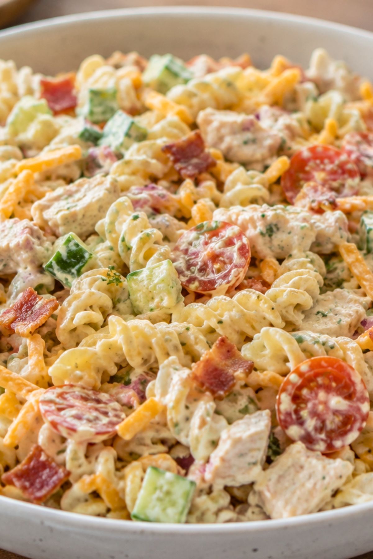 Creamy chicken bacon ranch pasta salad with rotini, grilled chicken, crispy bacon, cheddar, cucumber, cherry tomatoes, and ranch dressing in a white bowl.