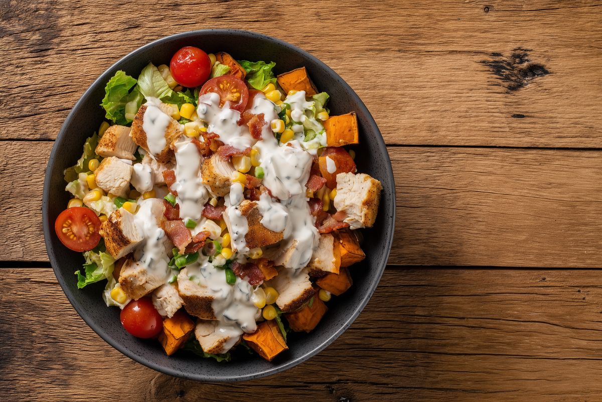 Chicken bacon ranch sweet potato protein bowl with roasted sweet potatoes, corn, tomatoes, lettuce, crispy bacon, and ranch dressing on a wooden table.