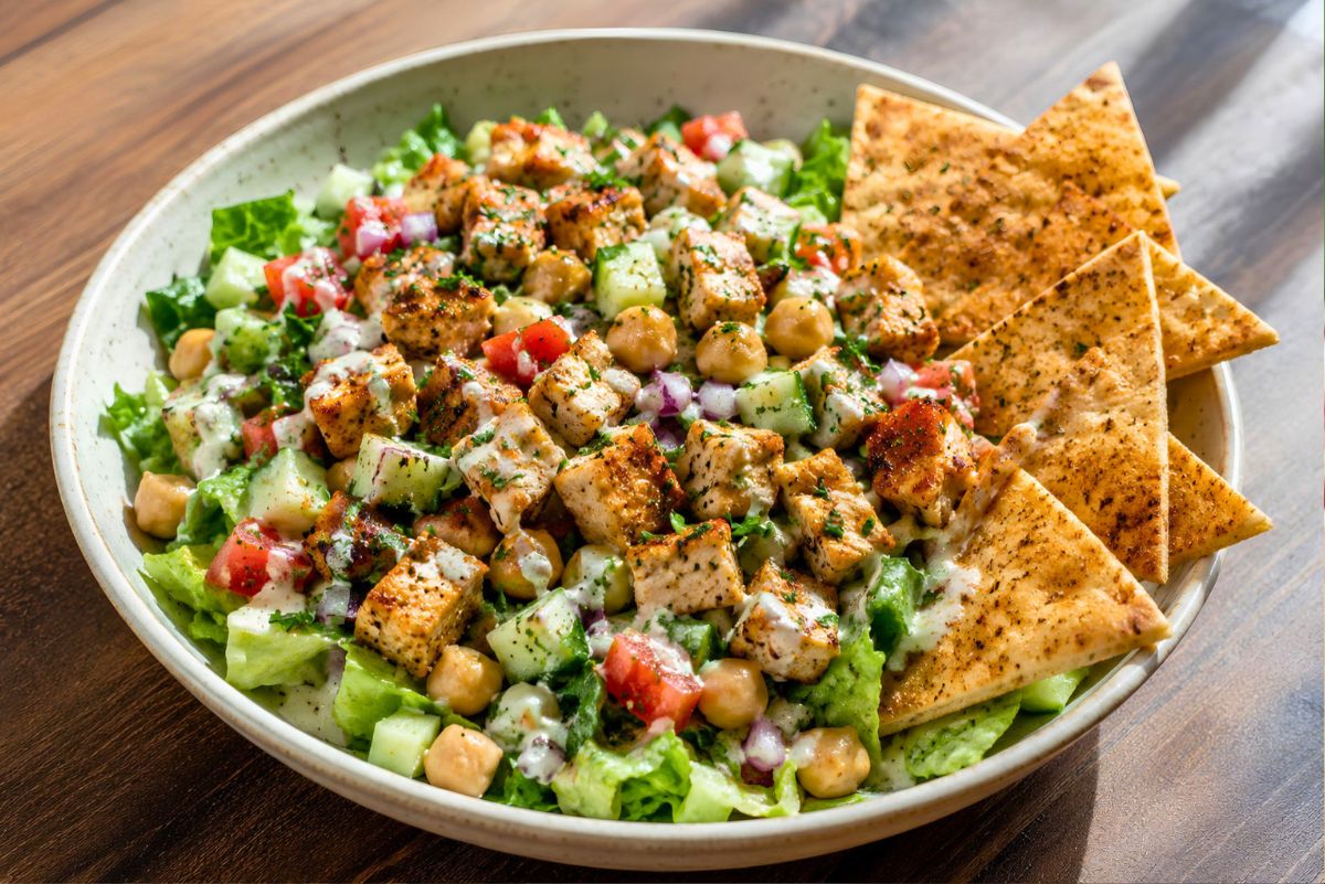 Chicken shawarma chopped salad with seasoned grilled chicken, chickpeas, cucumbers, tomatoes, red onion, romaine lettuce, creamy dressing, herbs, and toasted pita chips in a beige bowl.