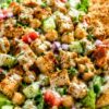 Chicken shawarma chopped salad with grilled chicken, chickpeas, cucumbers, tomatoes, red onion, romaine lettuce, creamy dressing, fresh herbs, and toasted pita chips in a beige bowl.
