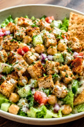 Chicken shawarma chopped salad with grilled chicken, chickpeas, cucumbers, tomatoes, red onion, romaine lettuce, creamy dressing, fresh herbs, and toasted pita chips in a beige bowl.