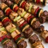Grilled chicken and steak kabobs on metal skewers with zucchini, red bell peppers, and scallions served on a white platter.