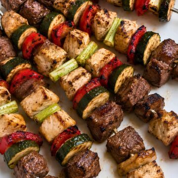 Grilled chicken and steak kabobs on metal skewers with zucchini, red bell peppers, and scallions served on a white platter.