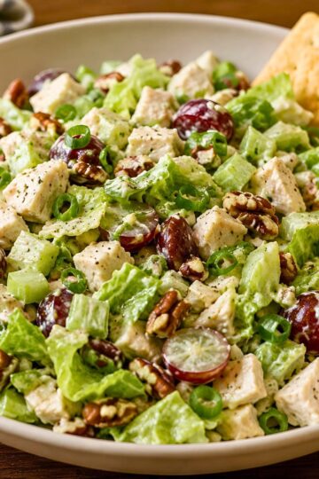 Chopped chicken salad with grapes, pecans, romaine lettuce, celery, green onions, and creamy dressing served in a white bowl with a cracker tucked in the back.