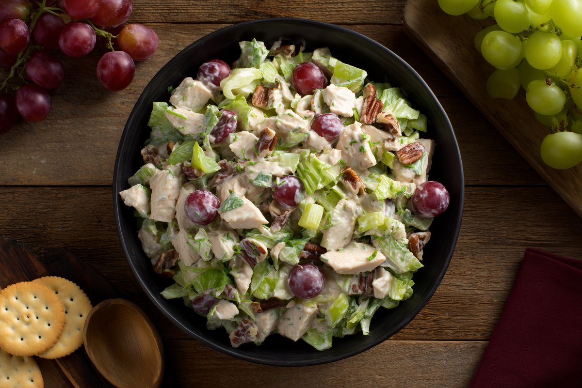 Chopped chicken salad with red grapes, pecans, celery, lettuce, and creamy dressing in a black bowl on a wooden table.