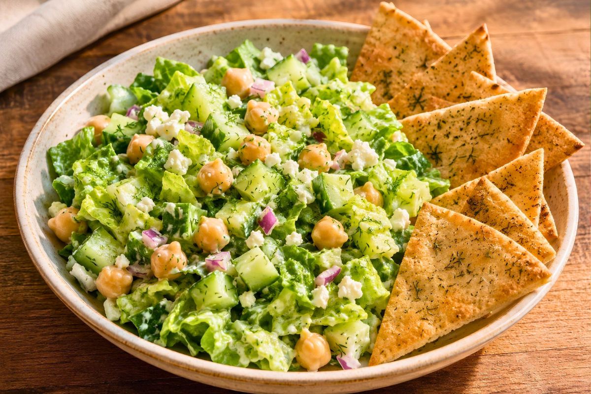 Creamy cucumber dill chickpea salad with romaine, cucumber, chickpeas, red onion, feta, fresh dill, and creamy dressing, served in a shallow bowl with toasted pita chips on a wooden table.