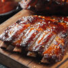 BBQ ribs with glossy sauce and sesame seeds on a wooden cutting board, with a sauce bowl and basting brush in the background.